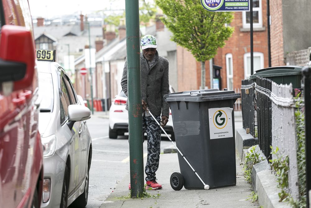 Man walking with a white cane, black bin in his way