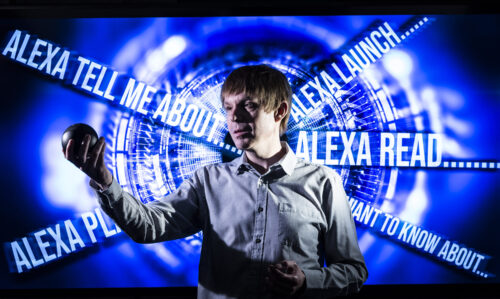 Robbie Ford holding a Smart Device in front of a screen containing Alexa Commands