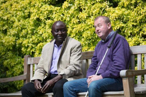 An NCBI service user who has a white cane sitting on a bench alongside another man