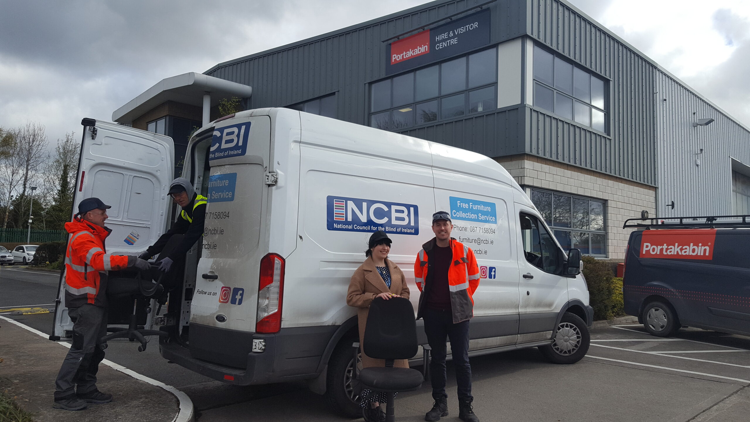 A representative from Portakabin and representatives from NCBI stand in front of a white NCBI van as they accept a donation of furniture from the company, some of which is being loaded into the back of the van