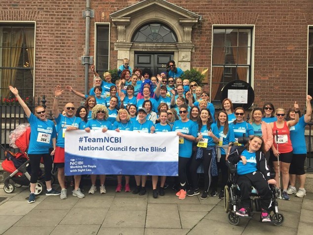 Team NCBI members pose together in a group with a banner at the VHI Women's Mini Marathon. Each member of the group is wearing a light blue t-shirt and the members at the front are holding a large banner to show they are Team NCBI.