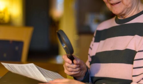 An older woman is reading a sheet of paper through a magnifying glass