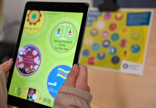 A close up of a child's hands holding a tablet. The child is using magnifying software on the tablet to read a yellow poster on a wall.