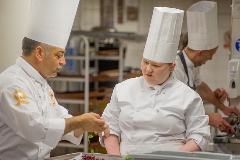 Chris Sandford is on the left and is wearing a chef's apron and hat. He is holding herbs and speaking to another female chef who is standing by his side.