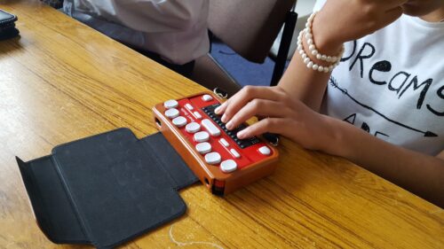 A young child is sitting beside a Braille technology trainer as they practice how to use a digital device which is read and also has white buttons.