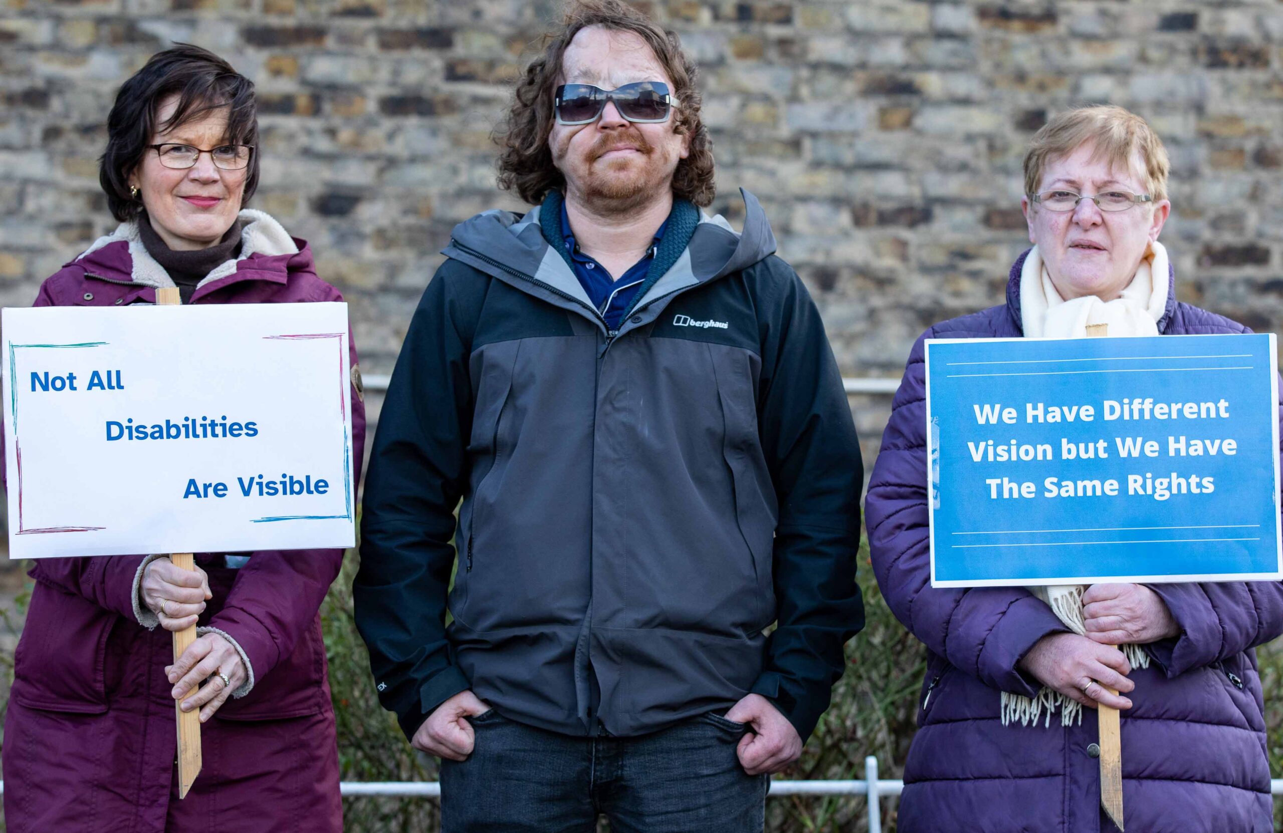 Three NCBI advocates standing together. There is a man in the middle and two women, one either side of him, who are holding signs. The sign on the left reads: Not all disabilities are visible. The sign on the right reads: We have different vision but we have the same rights.