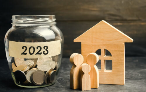A glass jar filled with coins to the left of the picture has a sticker on it which has the year 2023 visible. To the right of the jar are wooden figures of two adults and a child standing in front of a wooden house.