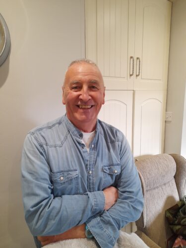 Eugene Rooney is smiling with his arms folded across his chest. He is wearing a light blue denim shirt.