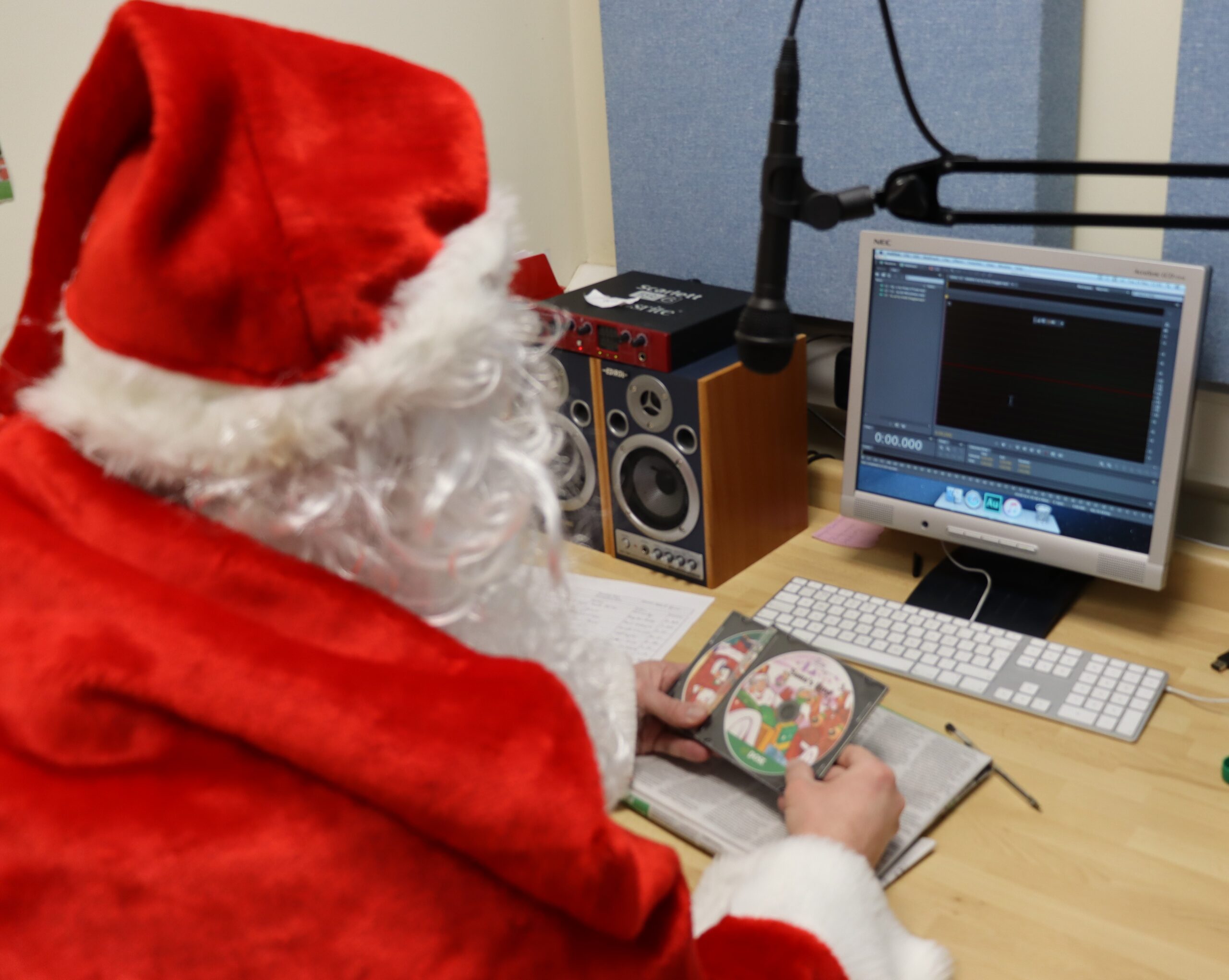 A picture of Santa sitting at a desk in front of a microphone and computer which has recording software on it. He is dressed in his red and white suit and is holding two audio CDs with letters for children who are blind or vision impaired.