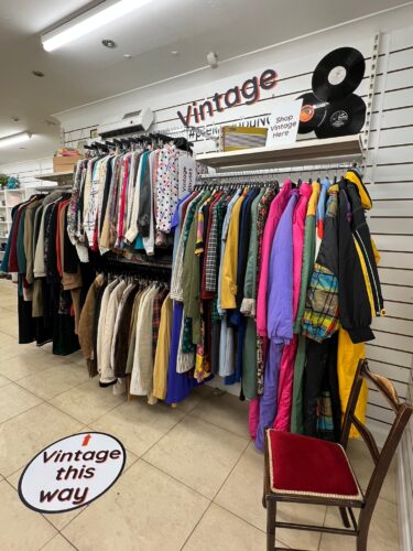 Rails of vintage clothing available in an NCBI store. There is a colourful selection available hanging from the wall, while vintage branding and old-style records decorate the area.