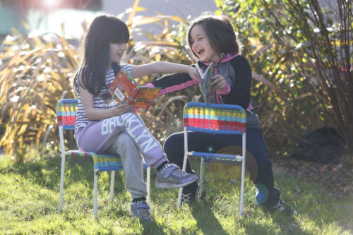 Two young girls are sitting on chairs on a patch of grass and they are both holding books. The girls are pointing at each others books and smiling. One child is wearing a purple and grey outfit and the other is wearing a black and pink outfit.