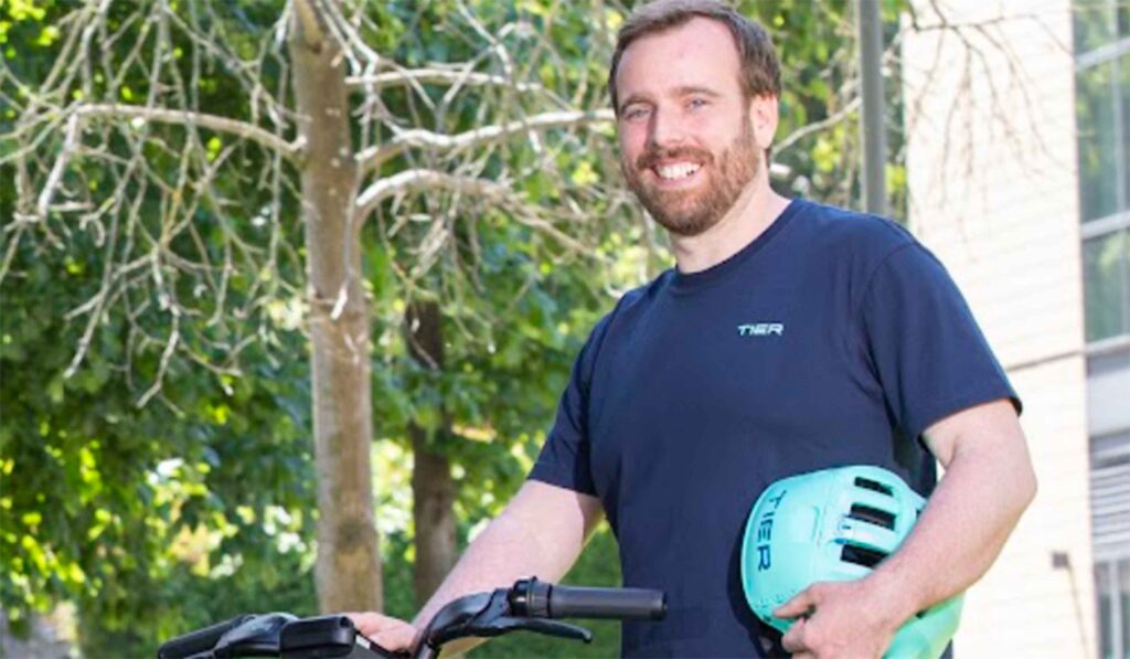 Peadar Golden is standing in a navy t-shirt and he is holding a bright green helmet in one arm and has his other hand resting on the handlebars of a a Tier e-scooter.