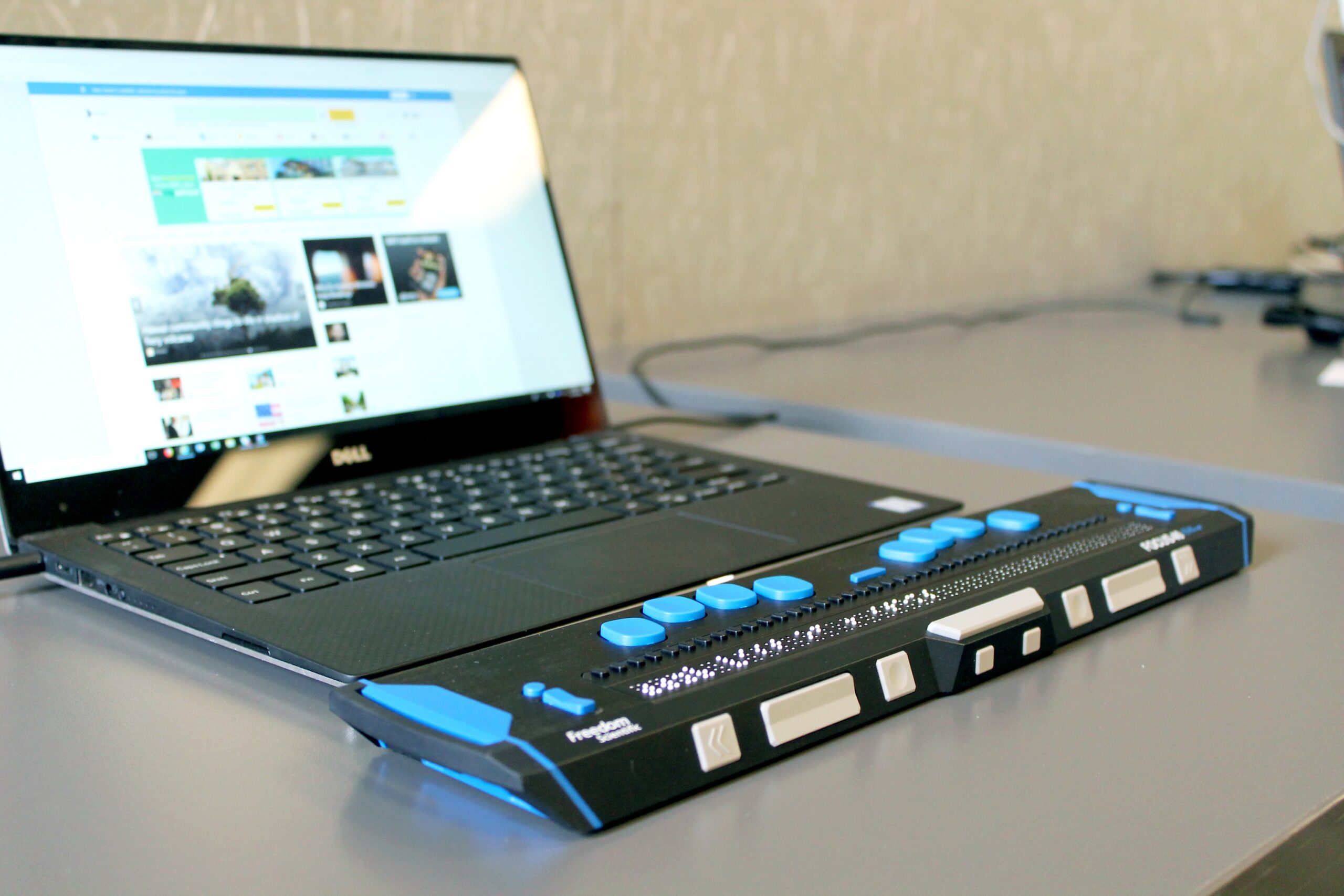 A digital Braille machine which is blue and black in colour is sitting in front of a laptop which is placed on a desk.