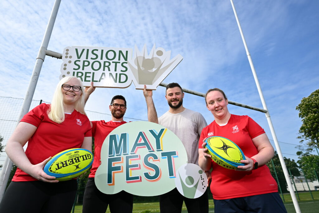 Two women in red tops are holding bright yellow rugby balls. In between them are Ian McKinley and Robbie Henshaw who are holding Vision Sports Ireland and Mayfest signs, respectively. All four people are standing under rugby goal posts.