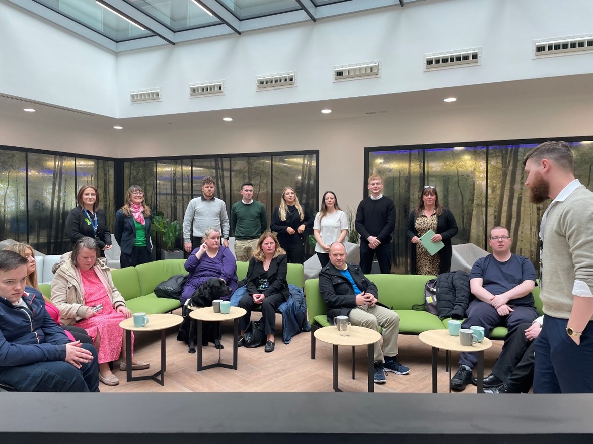 A group of NCBI service users are sitting around in a room on a green sofa as members of the CPL team are also gathered around while a person speaks at the front of the room.