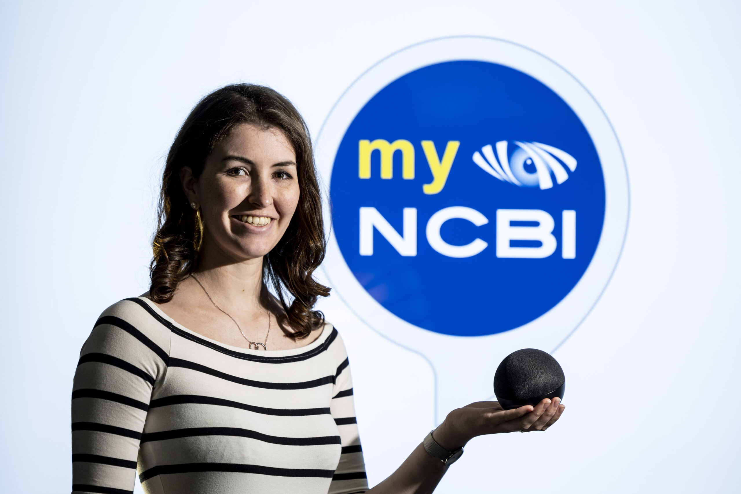 A young woman with dark hair is wearing a white top with black stripes. She is holding a smart speaker in front of the myNCBI Smart Hub logo, which is inside a blue circle.