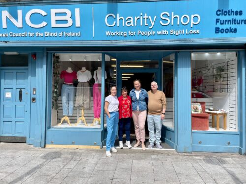 The NCBI Drogheda Team are huddled together and standing in the doorway of the store, which is painted light blue outside and has two windows with a clothing displays.