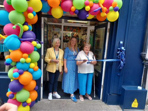 Store manager Jane Dwyer Hall, Carmel McDonnell and NCBI service user Helen Gray cutting the ribbon to open the store, under a multicoloured balloon arch.