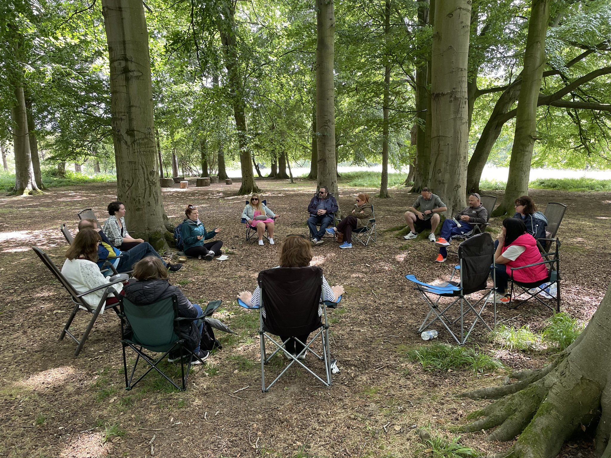 A group of participants in the Nature Connection Series sitting on camp chairs in a circle in an area of forest on one of the trips. They are surrounded by trees and are all facing each other in the circle.