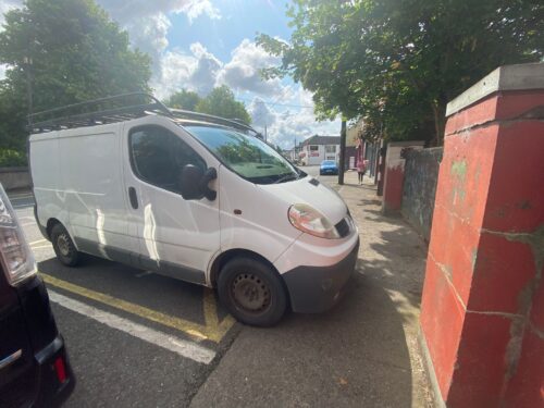 A white van is parked at the edge of a footpath. The front of the van is overhanging and taking up the majority of the space on the footpath, which is blocked by a wall on the other side.