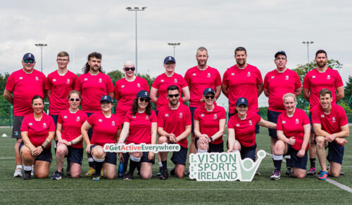 The Old Wesley squad posing for a group picture on a rugby pitch. They are all wearing the red and black kit for the team. They have a Vision Ireland sign in front of them.