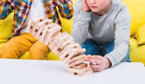 Two boys sitting on a yellow couch in front of a table. They are playing Jenga and the Jenga blocks are about to fall down. One boy is sad at losing and one is happy he won.